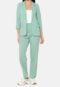 Miętowy garnitur z dopasowaną marynarką i luźnymi spodniami, wykonany z gładkiego materiału, zestawiony z białymi trampkami.