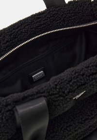 Czarna torba typu tote z teksturowanym, puszystym wykończeniem. Wewnątrz znajduje się kieszeń na zamek oraz mała metka z logo. W komplecie znajdują się syntetyczne uchwyty.