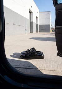 Sandales à plateforme noires avec boucles posées sur un sol en béton à l'extérieur d'un véhicule avec une porte ouverte dans une zone industrielle.