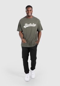 Camiseta color caqui con texto blanco "Smilodox Athlete", combinada con pantalones negros y zapatillas blancas. Mangas cortas y un ajuste relajado.