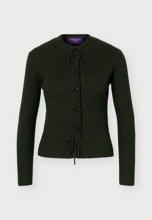 Un mannequin se tient devant un fond uni, vêtu d’un cardigan côtelé vert foncé, d’une longue jupe beige et de bottines noires à talons.