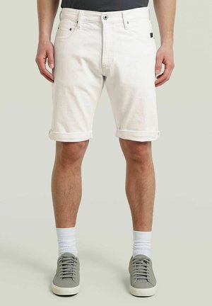 Mann trägt hochgekrempelte weiße Denim-Shorts, weiße Sportsocken und graue Turnschuhe, steht vor neutralem Hintergrund.
