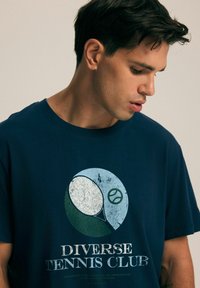 Granatowa koszulka z grafiką rakiety tenisowej i napisem "DIVERSE TENNIS CLUB" w białym i zielonym kolorze, z okrągłym wzorem kolorystycznym.