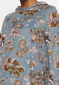 Mikina s kapucí v měkké modré látce s květinovým a listovým vzorem v růžových, oranžových a zelených tónech s texturovaným povrchem.