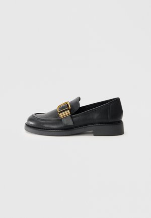 Black leather loafer with a flat sole, featuring a gold buckle accent and stitched detailing along the edges. Smooth texture.