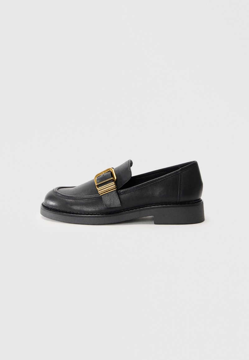 Black leather loafer with a flat sole, featuring a gold buckle accent and stitched detailing along the edges. Smooth texture.