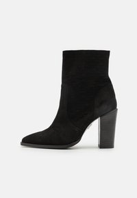 Botte cheville en daim noir avec bout pointu, dessus texturé et talon haut épais sur fond blanc.