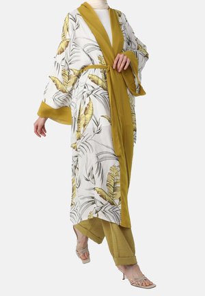 Femme portant une robe style kimono à motifs floraux blancs et jaunes avec des manches larges, associée à un pantalon moutarde et des talons beige à lanières.