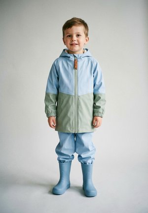 Young boy standing, wearing a two-tone blue and green raincoat with hood, matching blue rain pants, and blue rain boots on a plain background.