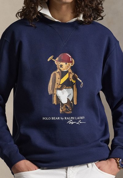 Temno modra jopa s kapuco z motivom medveda Polo, ki nosi rdečo kolesarsko čelado, karo jopico, jahalne škornje in drži polo palico. Napis se glasi "Polo Bear by Ralph Lauren."