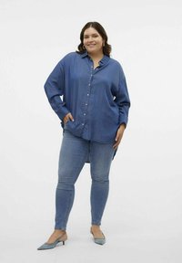 Camisa de mezclilla azul oscuro de botón, combinada con jeans ajustados azul claro y tacones azul claro. Presenta un corte relajado y mangas largas.
