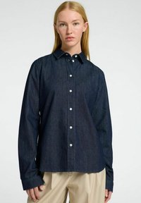 Tamsios denimo mygtukų marškiniai su apykakle, ilgomis rankovėmis ir baltais mygtukais. Medžiaga tekstūruota, parodanti subtilias vertikalias juostas.