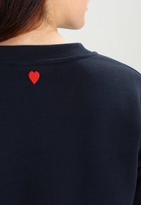 Sudadera azul marino con cuello acanalado, que presenta un pequeño corazón rojo bordado en la parte superior de la espalda. Textura suave, diseño casual.