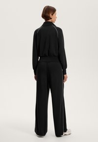 Fekete tréningruházat, cipzározott dzsekivel és széles szárú nadrággal, fehér díszítéssel. Az anyag lágy és laza megjelenésű.