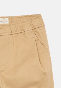 Brūni kokvilnas bikses ar elastīgu jostu, piedāvā sānu kabatas un gludu tekstūru. Ietver diegu šuvju detaļas.