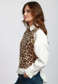 Gilet a stampa leopardata in beige e marrone, indossato sopra una camicia bianca con colletto, abbinato a jeans blu con dettagli a bottone.