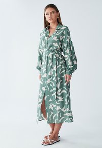 Zelena haljina s uzorkom, ovratnikom, dugim rukavima, prednjim gumbima, pojasom u struku i bočnim prorezom. Uparena s bijelim ravnim sandalama.