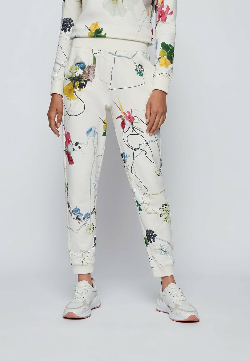 Blommönstrade sweatpants i vitt tyg, med flerfärgad botanisk tryck och elastiska muddar, i kombination med ljusa sneakers.