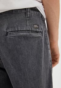 Šedé džínové kalhoty se zadní kapsou, na které je štítek s nápisem "Pull&Bear Original Denim Goods Established in 1991." Hladká textura.