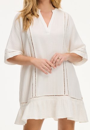 Femme portant une robe blanche ample et texturée avec des bandes découpées verticales, des manches mi-longues et un ourlet à volants, montrant plusieurs bagues en or aux doigts.