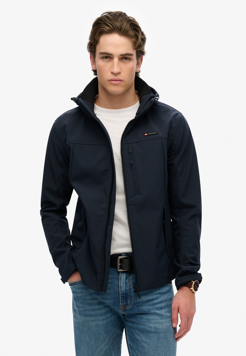 Giacca softshell blu navy con tasca sul petto con cerniera, cappuccio regolabile, colletto alto e interno testurizzato, indossata sopra una maglietta bianca.