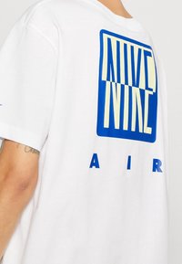 Vit t-shirt i bomull med en stor blå och gul grafisk text som läser "NIKE NINE AIR" på baksidan, med rund hals och korta ärmar.
