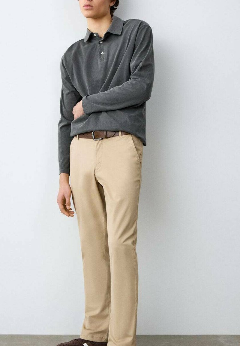 Persoană tânără care poartă un tricou polo cu mânecă lungă gri închis, pantaloni bej, curea maro și pantofi maro închis, stând în picioare în fața unui perete simplu, deschis la culoare.