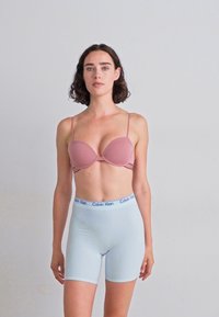 La modella si erge con sicurezza, indossando un reggiseno rosa pallido e pantaloni corti blu Calvin Klein, su uno sfondo grigio semplice.