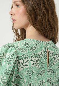 Blouse verte à motif floral avec manches courtes bouffantes et fermeture dos en forme de goutte, en tissu texturé avec des motifs de feuilles détaillés.