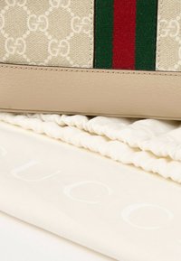 Sac en cuir beige et toile monogrammée GG avec bande verte et rouge posé sur un tissu blanc avec logo Gucci visible.