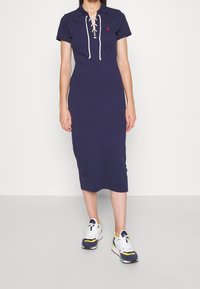 Žena oblečená v tmavě modrých midi šatech s krátkým rukávem a šněrováním, v kombinaci s multikolorovými bílými teniskami, stojící na jednobarevném pozadí.