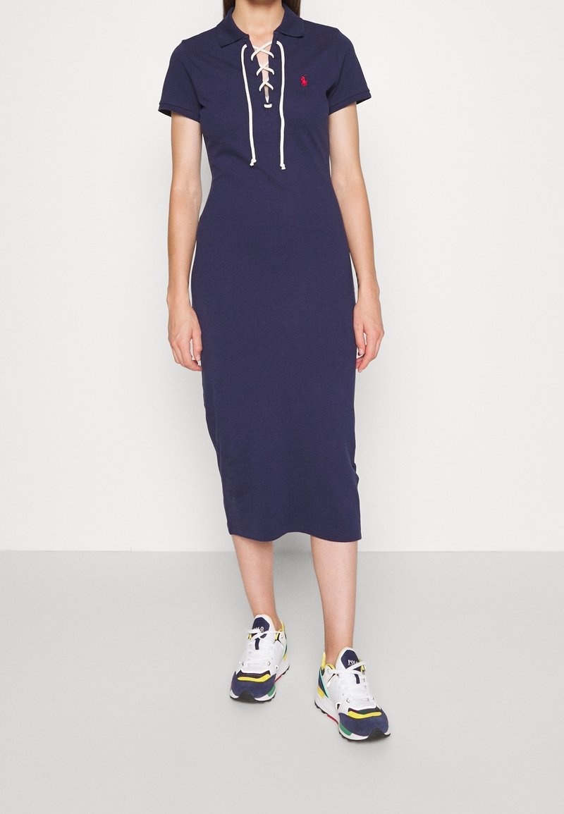 Žena oblečená v tmavě modrých midi šatech s krátkým rukávem a šněrováním, v kombinaci s multikolorovými bílými teniskami, stojící na jednobarevném pozadí.