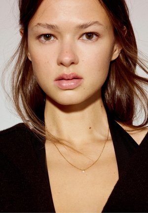 Jeune femme aux cheveux bruns, portant un délicat collier en or et un haut noir, face à l'avant avec une expression neutre sur un fond uni.