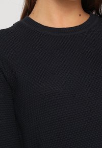 Tmavě modrý pletený svetr s texturovaným vzorem, širokým kulatým výstřihem a dlouhými rukávy. Tkanina působí měkce a mírně pružně.