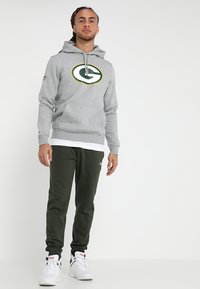 Sudadera gris con un logo verde y amarillo, con un bolsillo frontal, combinada con pantalones de chándal verde oscuro y zapatillas blancas.
