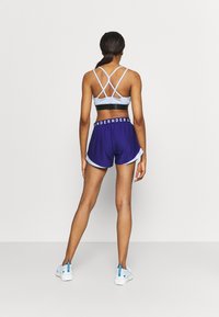 Světlomodrá sportovní podprsenka s křížovým zadním designem a černým páskem, kombinovaná s fialovými sportovními šortkami s bílým lemem a páskem s logem.