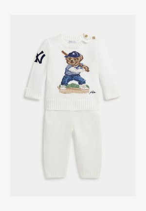 Weißes gestricktes Babyoutfit mit langen Ärmeln, auf der Vorderseite ist ein Bär in Baseballausrüstung gestickt und auf dem Ärmel befindet sich ein dunkelblaues NY-Logo.