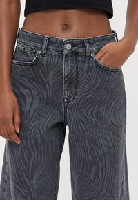 Svart jeans med et bølgete zebra-mønster, høy midje, fork pocket og metallknapplukking. Stoffet har en lett glans.