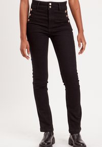 Jean en denim noir taille haute avec des accents de boutons sur les côtés et une coupe ajustée, associés à des bottines noires.