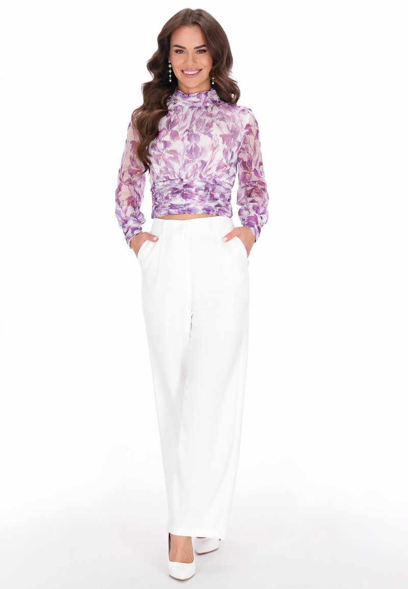 Vrouwelijk model in een lavendelbloemige doorschijnende blouse met hoge hals en witte wijde broek. Bevat zakken en eenvoudige witte hakken.