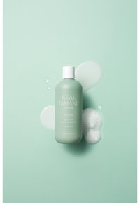Grön plastflaska med schampo och ett vitt lock, märkt "Real Tamanu." Innehåller stänk av klar vätska och skum mot en myntgröna bakgrund.