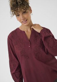 Bordeaux bluse med detaljeret blomsterbroderi på forsiden og ærmerne, med en V-hals, to knapper og lange, let puffede ærmer.