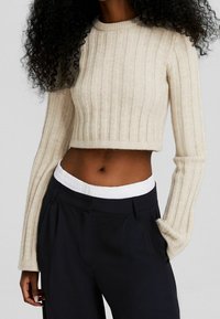 Beige ribbad kortärmad tröja med lång ärm och rund halsringning, tillsammans med mörka byxor med ett vitt elastiskt midjeband.