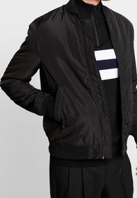 Blouson aviateur noir à la texture lisse et brillante. Comprend une fermeture éclair à l'avant, des poignets côtelés et des poches latérales. Porté sur un pull rayé noir et blanc.