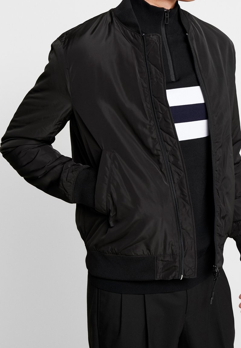Blouson aviateur noir à la texture lisse et brillante. Comprend une fermeture éclair à l'avant, des poignets côtelés et des poches latérales. Porté sur un pull rayé noir et blanc.