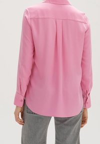 Personne portant un chemisier rose à manches longues avec un pli au dos et un pantalon gris, vue de dos sur un fond neutre.