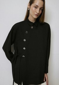 Zwarte lange mouwen shirt met een gestructureerde kraag, featuring grote metalen knopen en een gestrikte taille detail, gemaakt van een gladde stof.