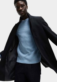 Sort frakke med høj krave, båret over en lyseblå sweater. Frakken har en glat overflade og store frontlommer.