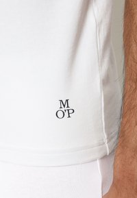 T-shirt in cotone bianco con maniche corte, caratterizzato da un piccolo logo nero "M O'P" vicino all'orlo, texture liscia e design minimalista.