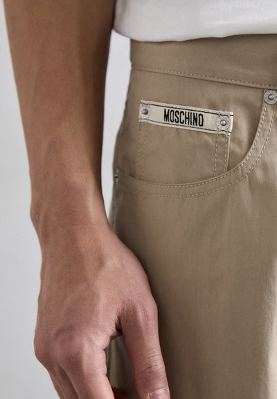 Bežiniai kelnės su matomu Moschino ženklu, tvirtu siuvimu ir metalinėmis detalėmis. Lygi tekstūra su prigludusia modeliacija.